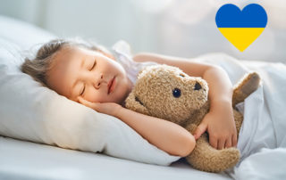 Kind schlafend im Bett mit Teddybär, oben Rechts ein Herz in den Farben der Ukraine